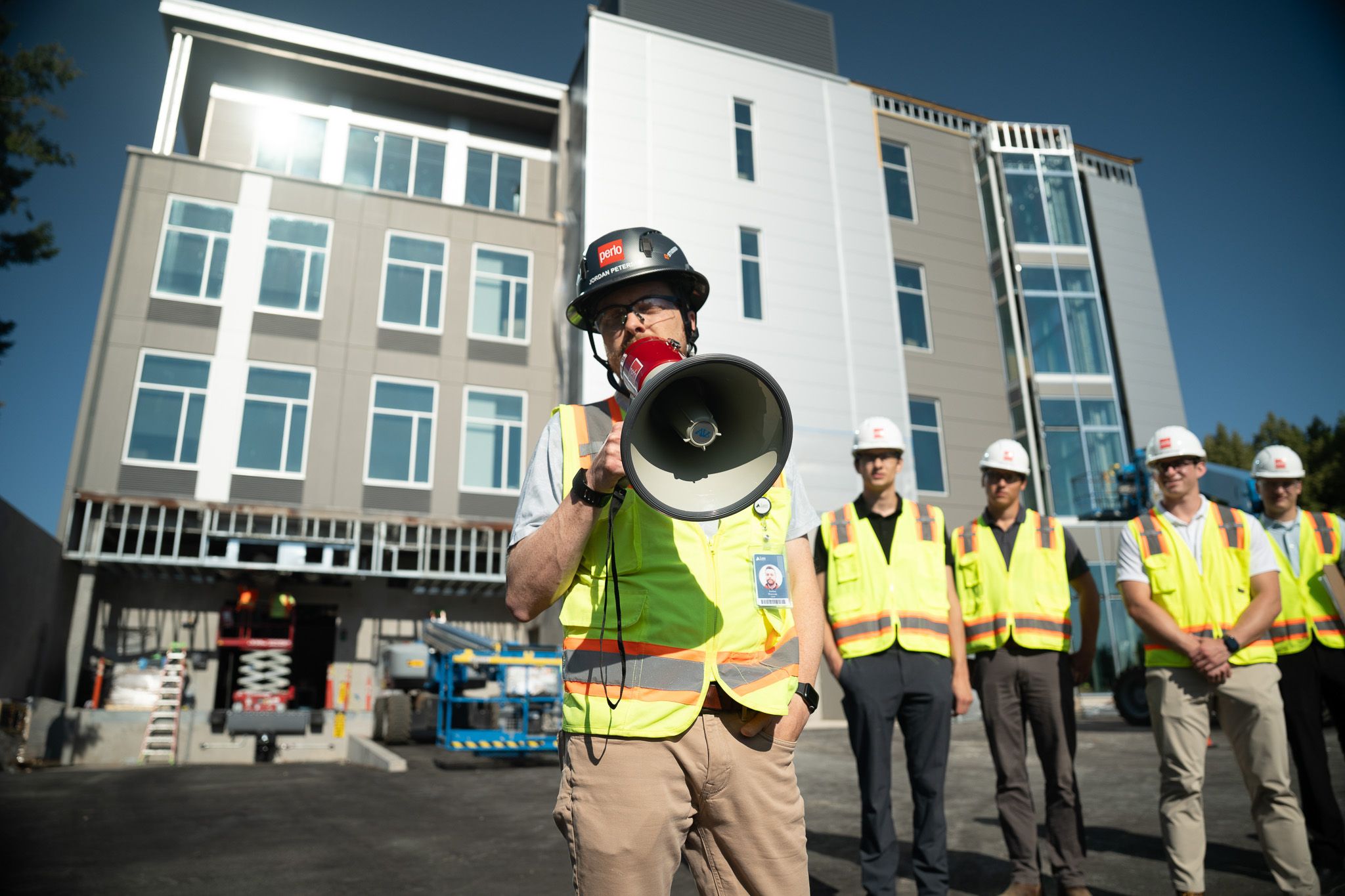Man speaks into a megaphone standing in front of an office building wearing a hard hat and high visibility vest