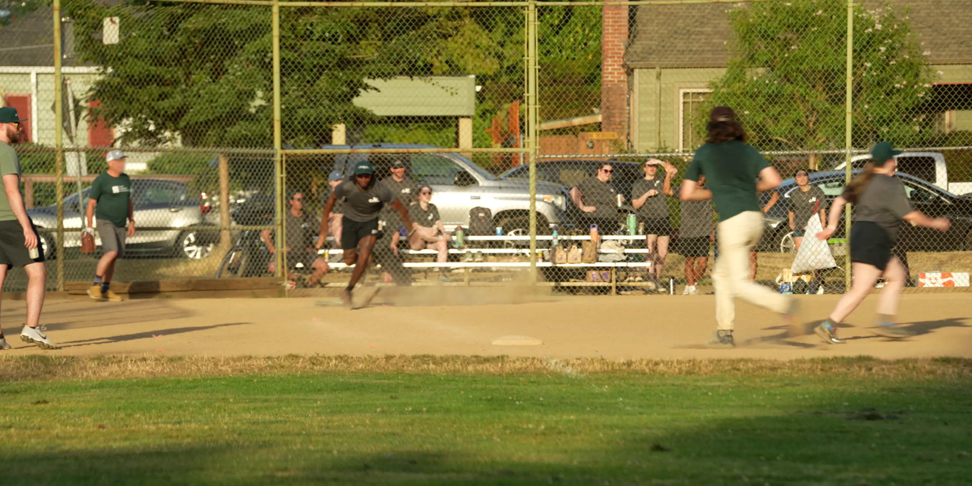 Softball Team in Woodstock Park in Portland. A batter is running to first base while fielders are reacting to his hit.
